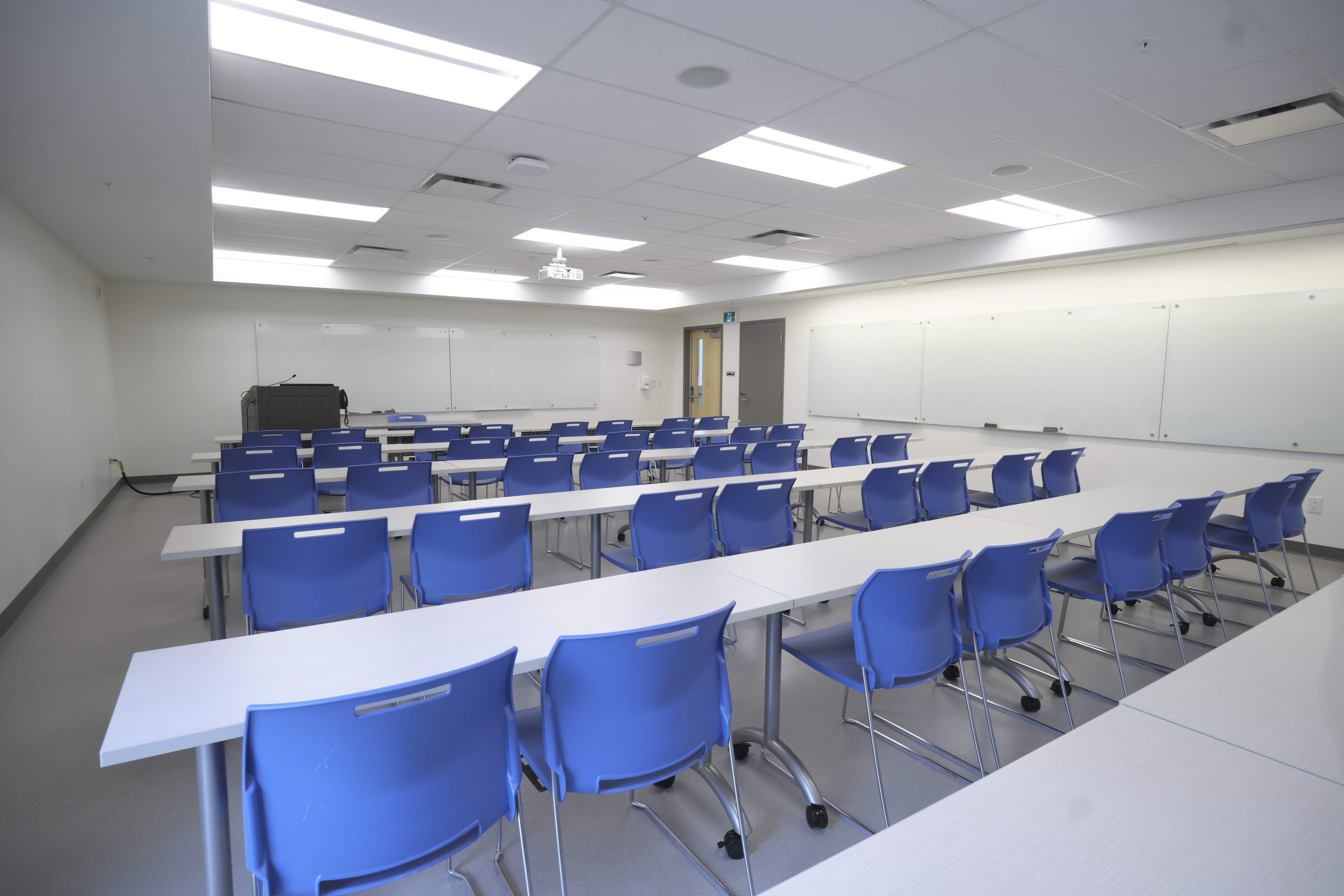 A room with rows of tables and chairs with a lectern at the front and whiteboards along the front and side wall.