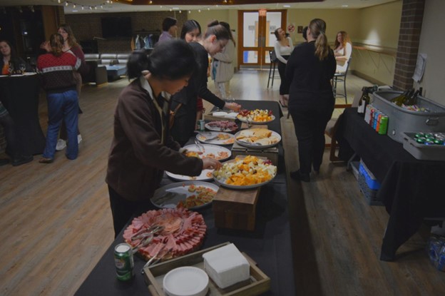 Students serve themselves from a buffet spread in the foreground as other students in the background sit or stand around cocktail tables chatting and laughing.