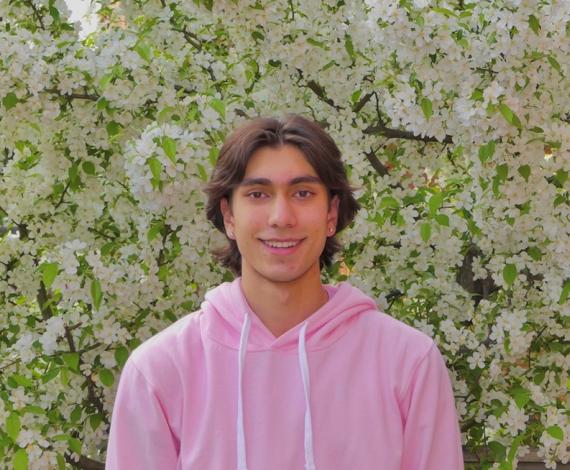 Asher, a masc presenting person, smiles broadly in front of a flowering bush.